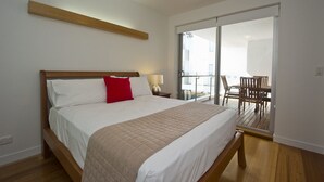 Un dormitorio con una cama grande, cabecera de madera, almohada roja, lámpara de noche y puerta corrediza de vidrio que da a un balcón con mobiliario exterior.