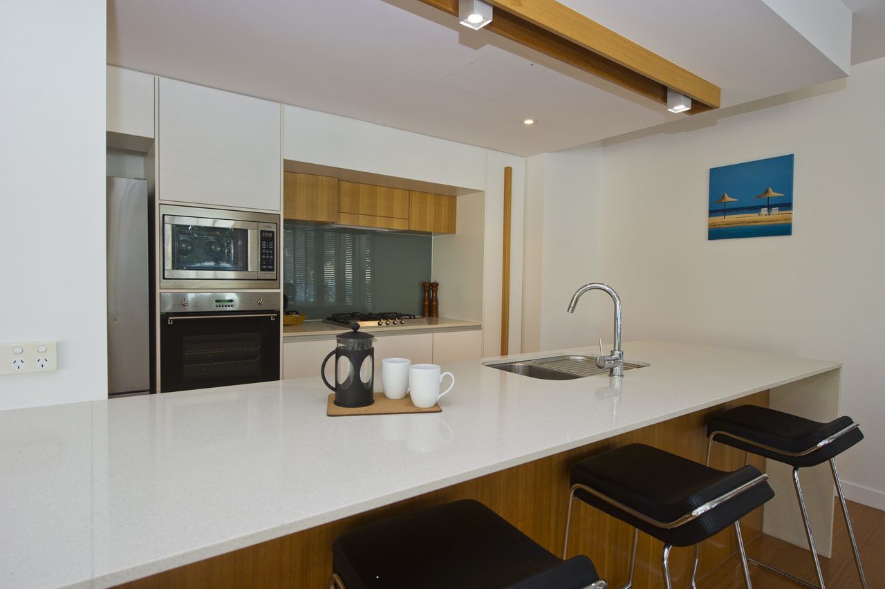 A modern kitchen with a built-in oven, microwave, and a coffee maker on the counter. There are two black bar stools and a sink with a faucet.