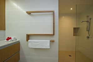 A bathroom with white tiled walls, a wooden towel rack, a glass shower enclosure, and a wooden vanity with a towel and red flowers.