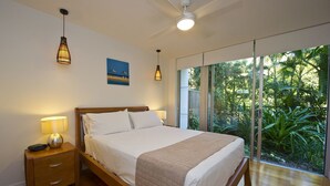 Quarto com cama de madeira, abajures, mesa de cabeceira, uma janela grande com vista para o verde e um ventilador de teto.