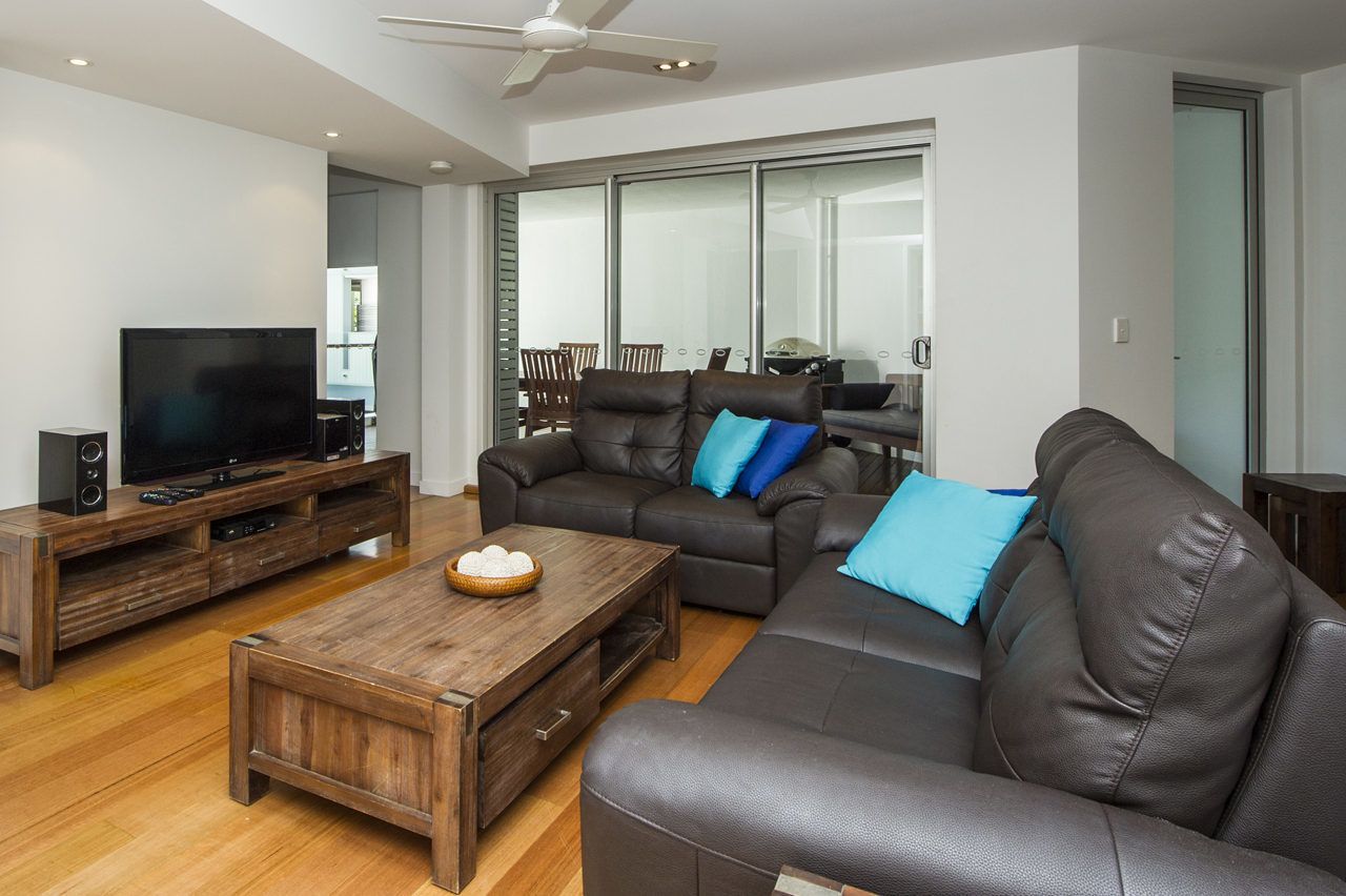 A modern living room with a large flat-screen TV, leather sofas, a wooden coffee table, and a ceiling fan.