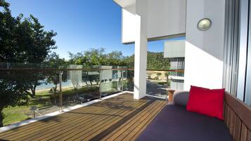A balcony with a wooden floor, a bed with a red pillow, and a glass railing overlooking a body of water and greenery.