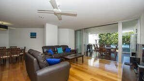 A modern living room with a sofa, coffee table, and dining area. There is a ceiling fan, a painting on the wall, and a sliding glass door leading to an outdoor patio. - Maggies Beachfront Apt. 7 (Horseshoe Bay)