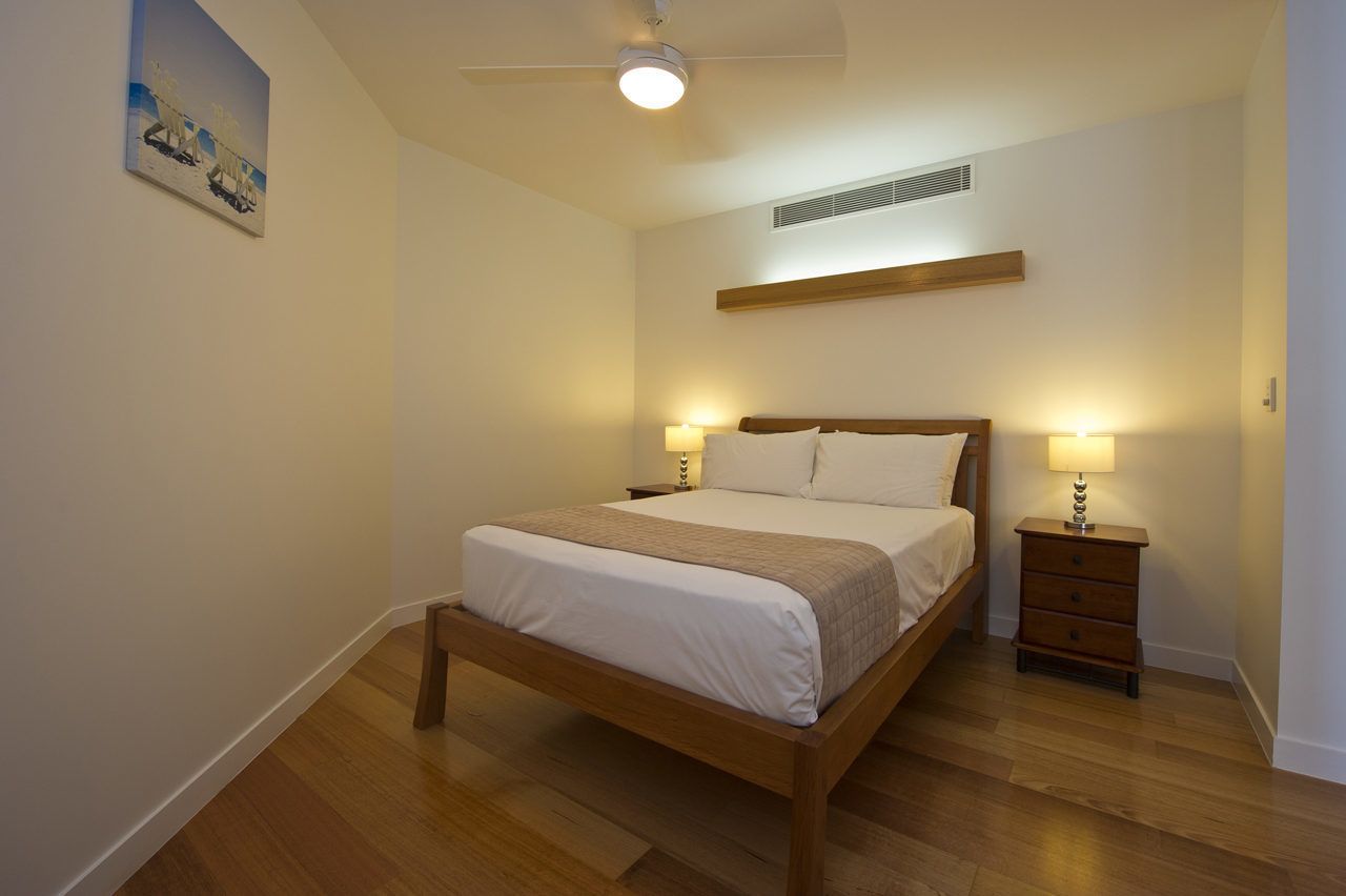 A bedroom with a wooden bed, two bedside tables with lamps, a ceiling fan, and a painting on the wall.