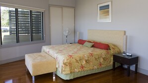 Une chambre à coucher avec un grand lit, une table de chevet, un ventilateur sur pied et une fenêtre avec des stores.