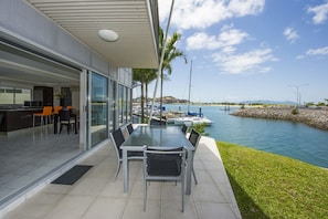 Un área de comedor exterior moderna con mesa y sillas de vidrio, con vista a un puerto deportivo con barcos y una masa de agua tranquila.