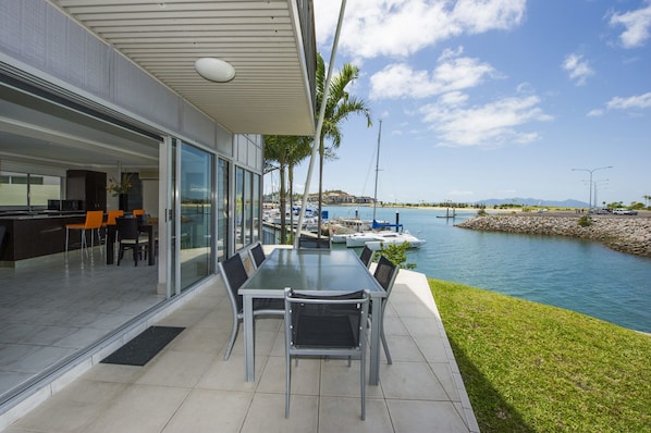 A modern outdoor dining area with a glass table and chairs, overlooking a marina with boats and a calm body of water.