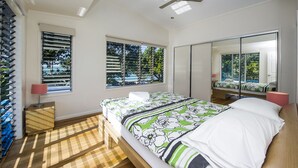 Une chambre à coucher avec un lit, deux fenêtres équipées de stores, un ventilateur de plafond, un miroir et un plancher en bois.