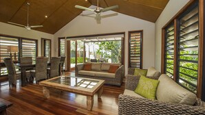 A spacious living room with a wooden ceiling, a ceiling fan, a glass coffee table, wicker furniture, and large windows with shutters. - Pure Magnetic Villa 1 (Nelly Bay)