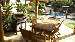 An outdoor patio with a thatched roof, a grill, a dining table with chairs, and a view of a swimming pool and greenery.