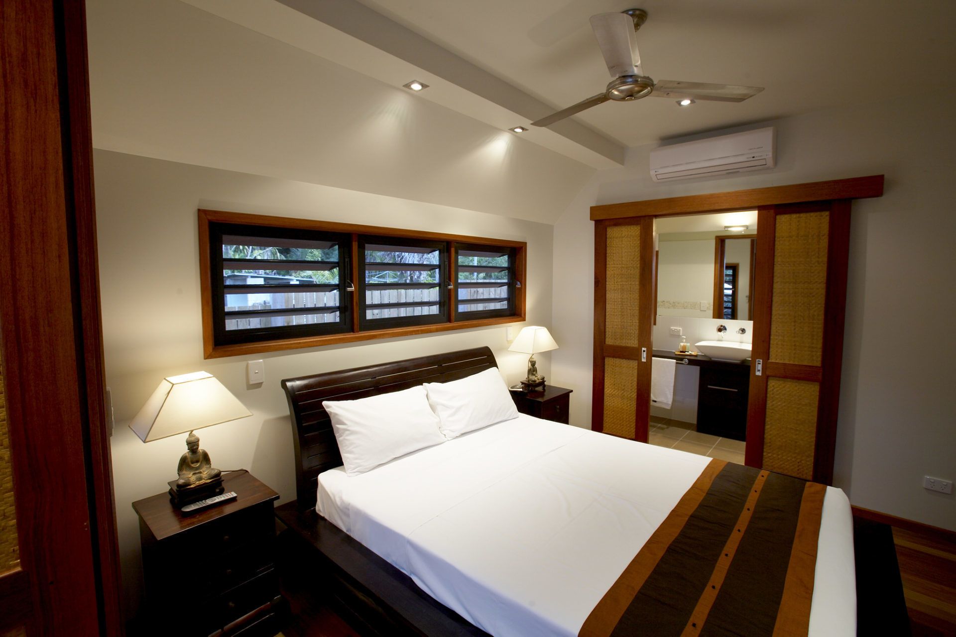 A bedroom with a bed, two bedside lamps, a ceiling fan, and a window with a view of a wooden deck.
