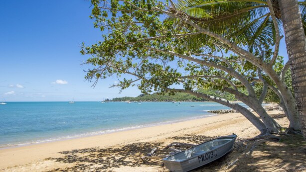 Une plage avec un bateau nommé « M1073Q » à l’ombre d’un arbre, une eau d’un bleu limpide et des bateaux visibles au loin.