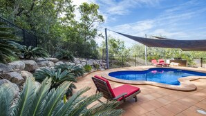 Une piscine à bord incurvé, entourée d’une terrasse carrelée, d’un fauteuil de relaxation rouge et d’un espace ombragé abrité par un auvent.