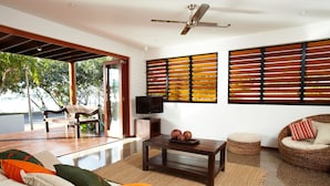 A modern living room with a sofa, coffee table, and a television. There are large windows with blinds, a ceiling fan, and a bench outside. - Villa Kembali (Horseshoe Bay)