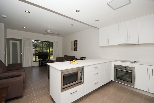 A modern kitchen with white cabinets, a built-in oven, and a microwave. There is a dining area with a table and chairs, and a sofa in the living room.