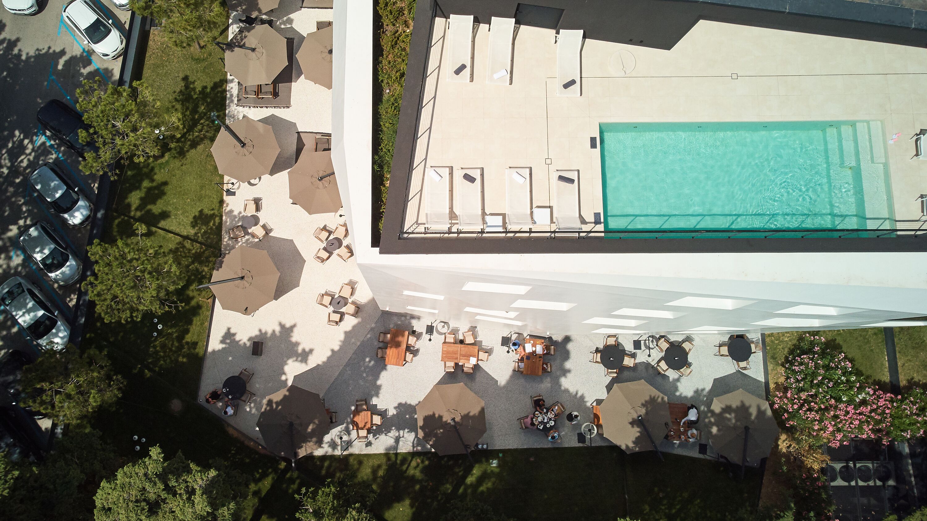 An aerial view of a rooftop pool area with multiple lounge chairs and umbrellas, surrounded by trees and a parking lot.