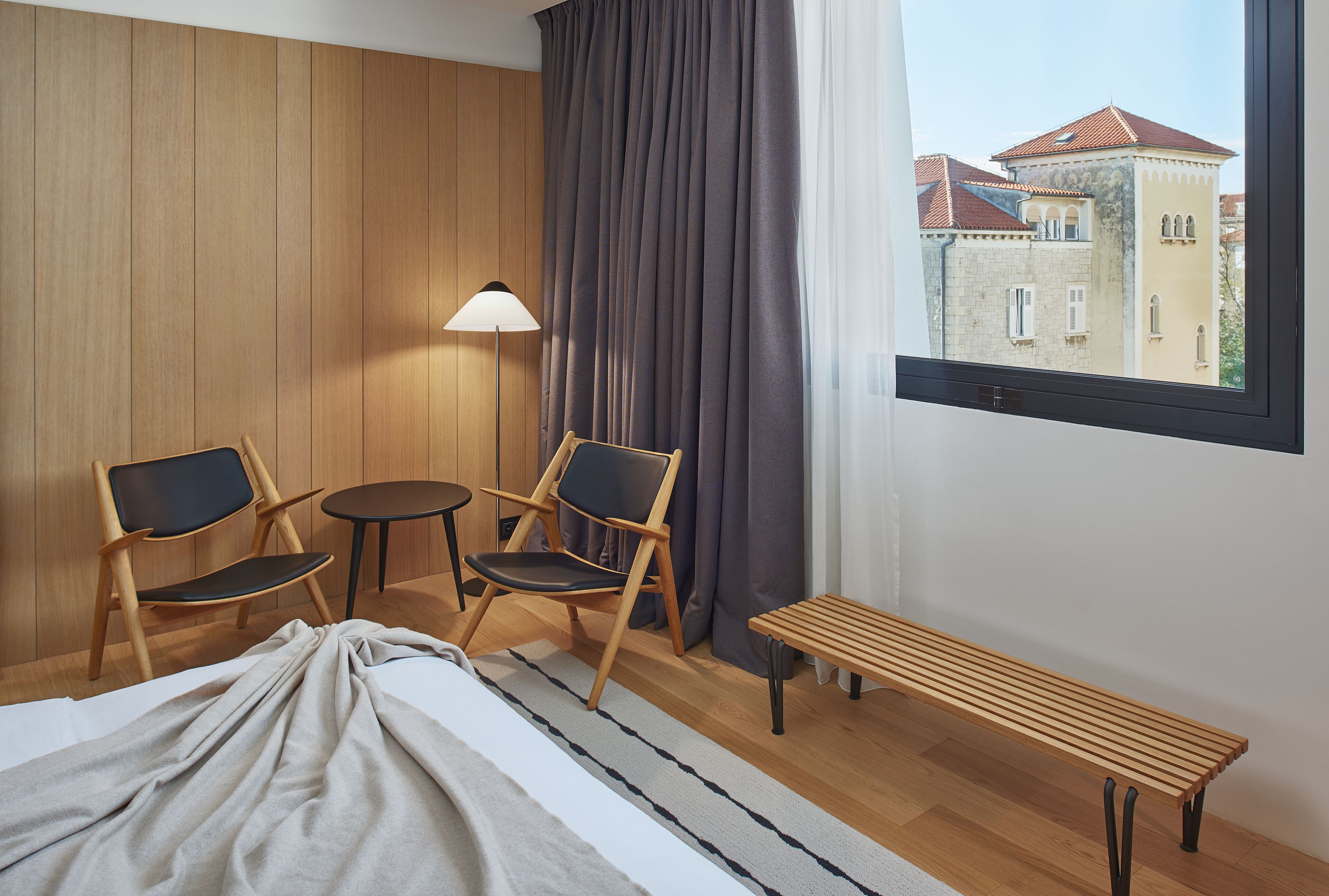Habitación de hotel con cama, dos sillas, una mesita, un banco, una lámpara y ventana con vista a un edificio.