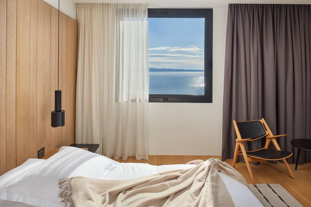 A bedroom with a large window, a bed, a chair, and a view of the sea.