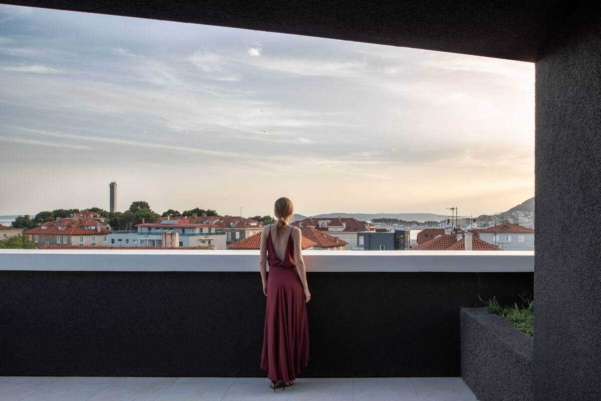 A woman in a long dress standing on a balcony overlooking a cityscape with red-roofed buildings and a tall tower in the distance.