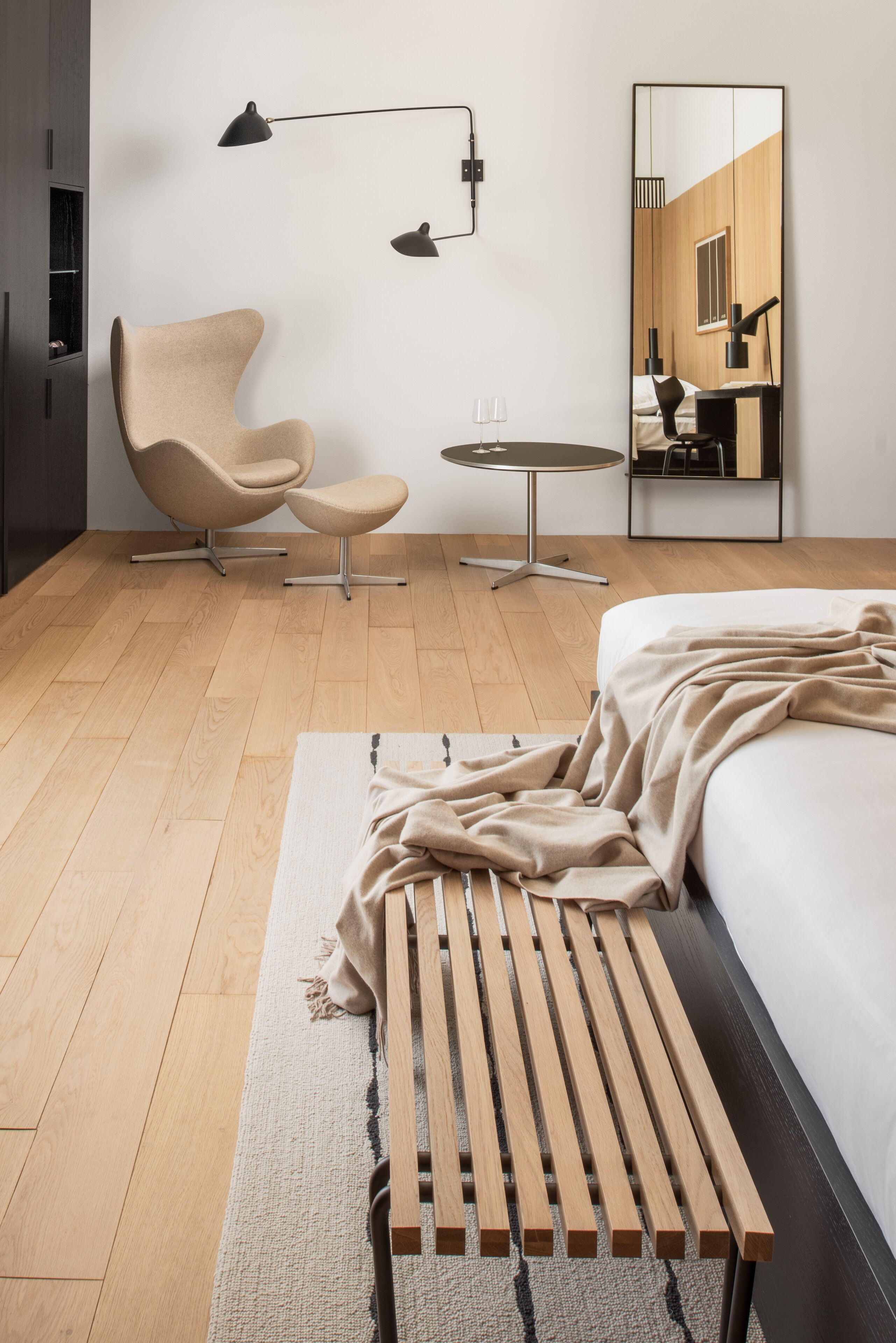 A modern bedroom with a wooden floor, a large bed, a chair, a small round table, and two floor lamps.