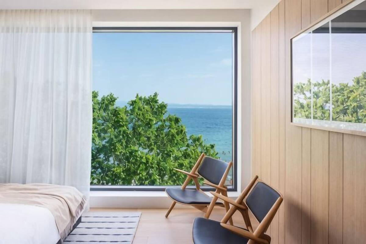 A bedroom with a large window, a bed, a chair, and a view of the sea.