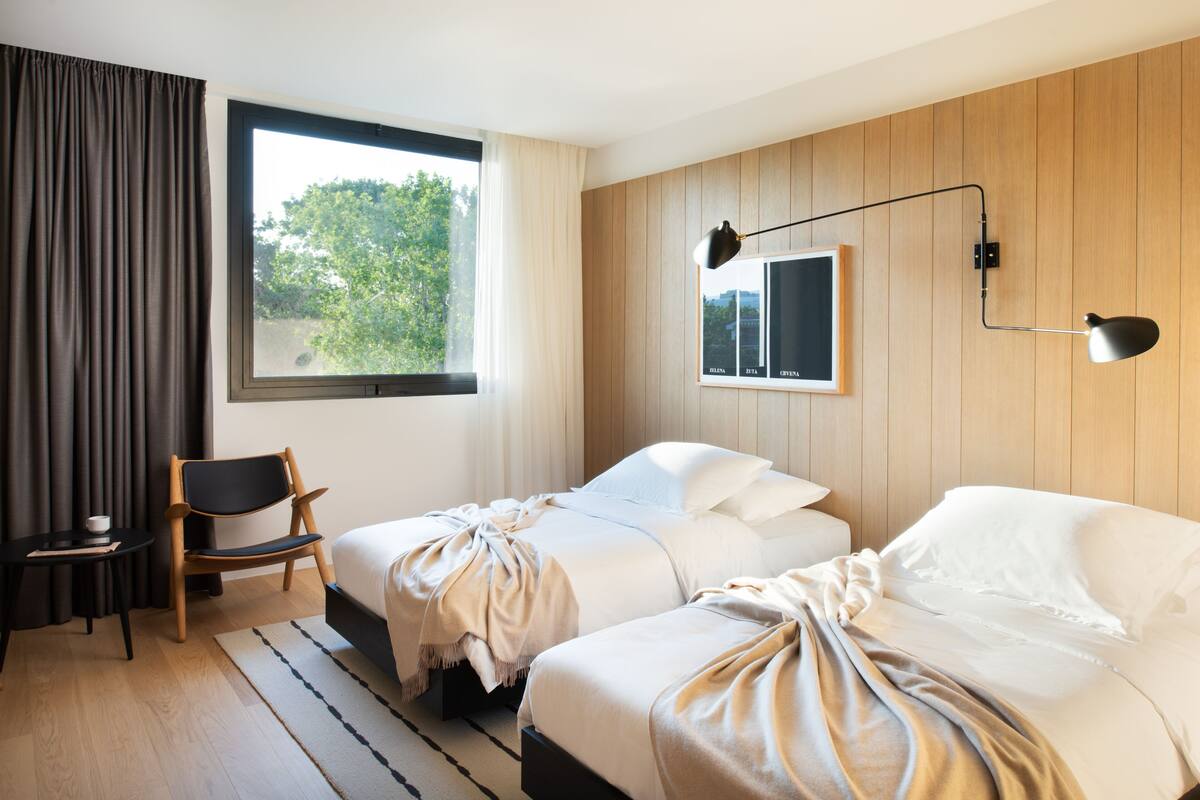 A hotel room with two beds, a wooden wall panel, a window with curtains, a desk with a chair, and a wall-mounted lamp.