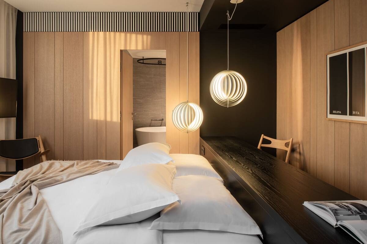 A modern hotel room with a bed, a wooden dining table, and pendant lights.