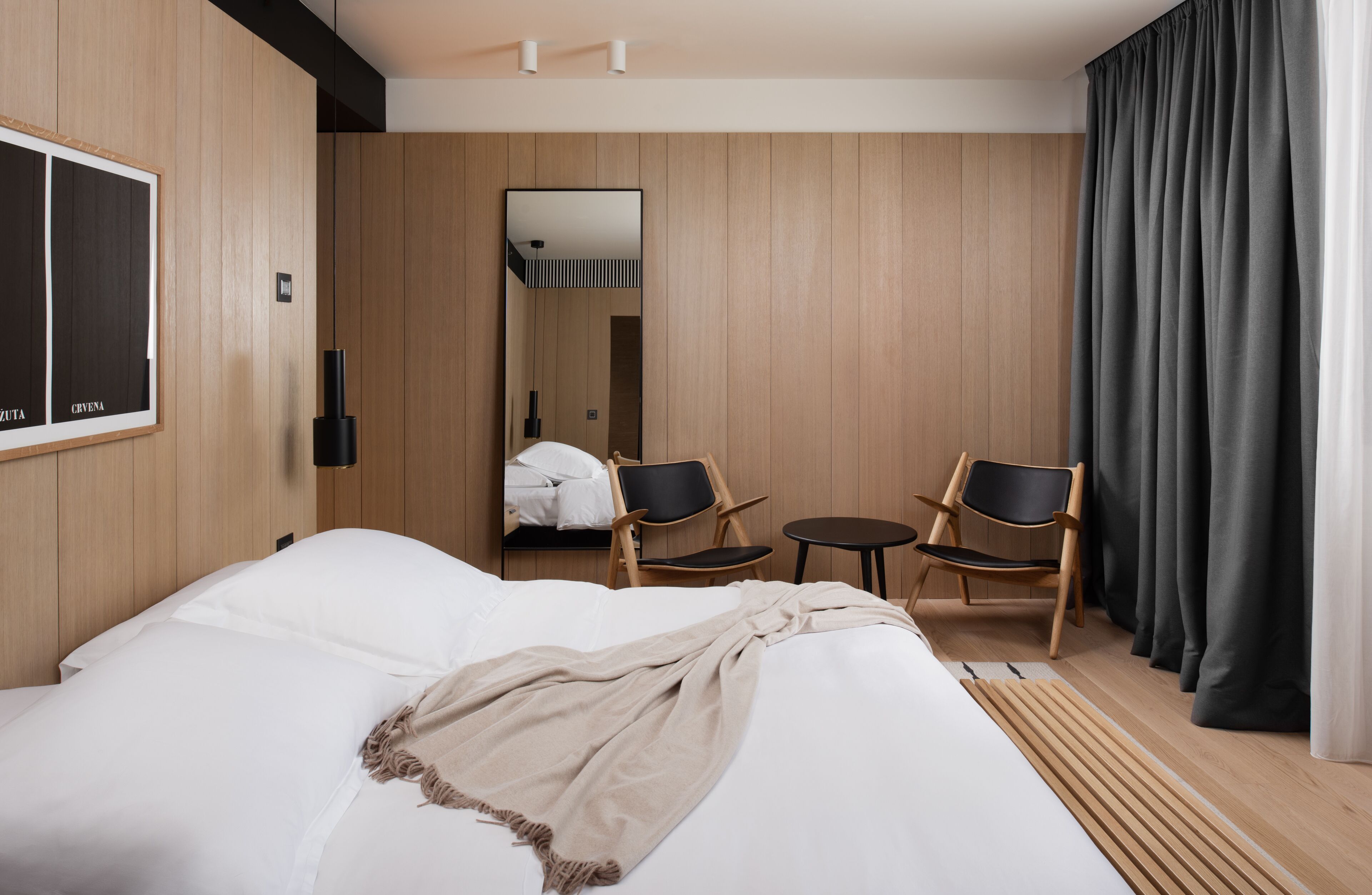 Une chambre d’hôtel moderne avec un grand lit, deux fauteuils, une petite table, un miroir et un lambris en bois.