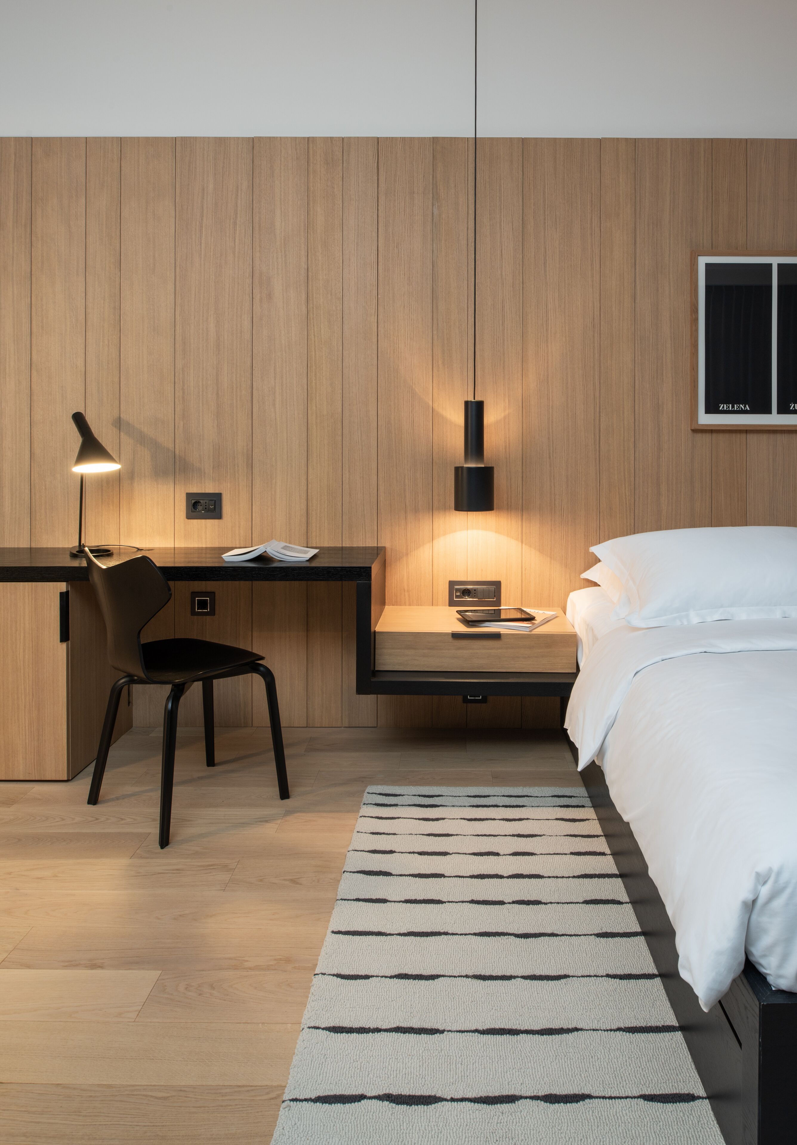 A modern bedroom with a wooden wall, a desk with a chair, a bed, and a nightstand.