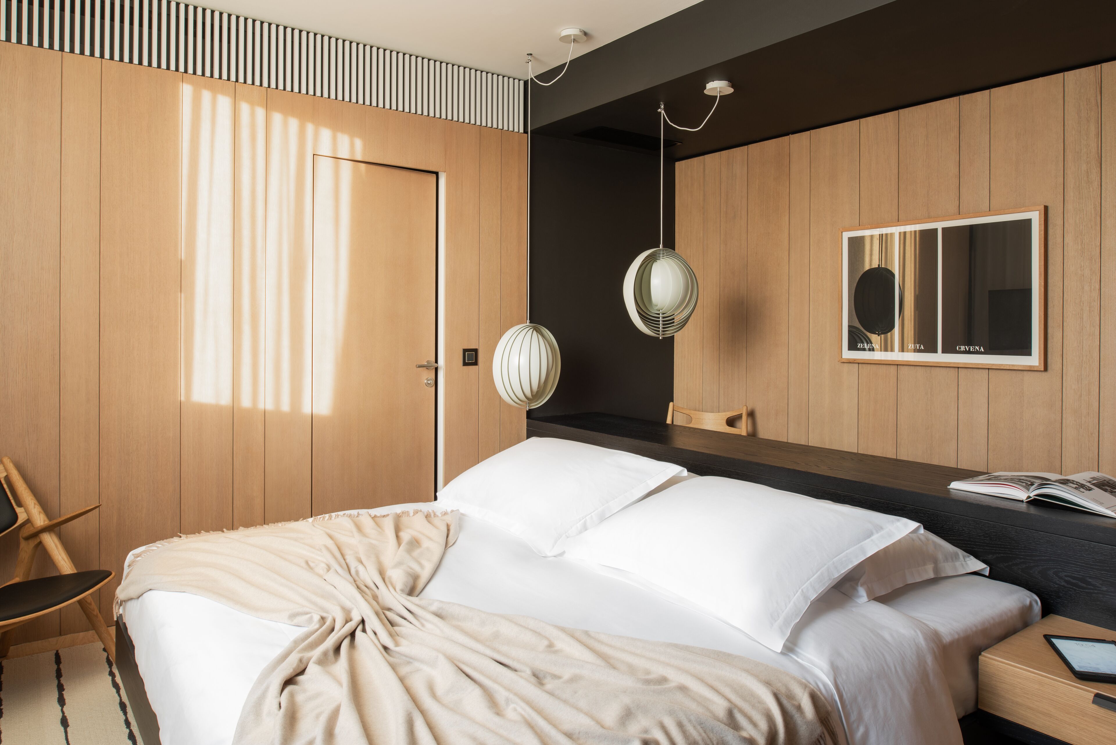 A modern bedroom with a large bed, wooden panel walls, a bedside table with a book, and a hanging light fixture.