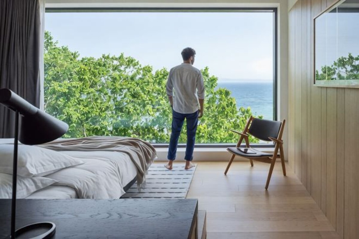 Un dormitorio con una ventana grande, una cama, una silla y vistas a la vegetación y al mar.