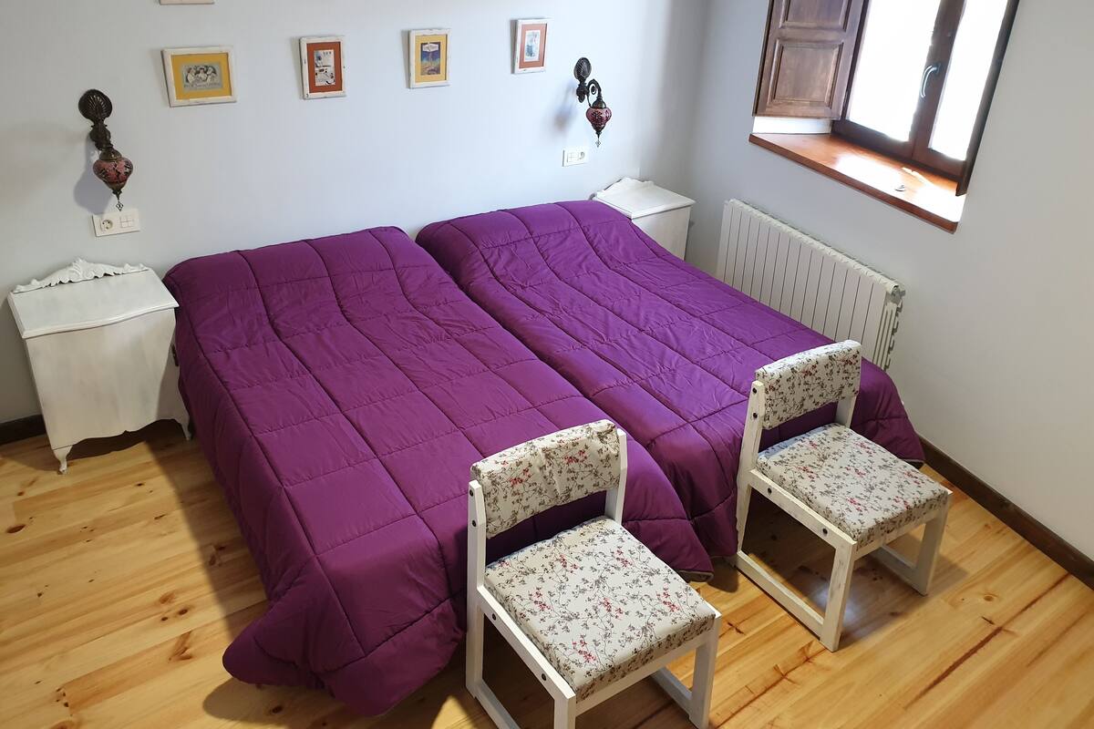 A room with two beds, a window, a radiator, and a wall with framed pictures.