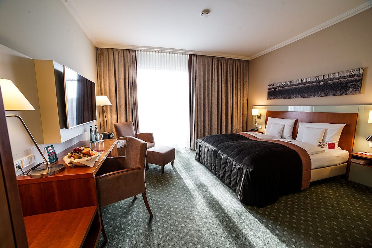 A hotel room with a bed, a desk with a chair, a TV, and a large window with curtains.