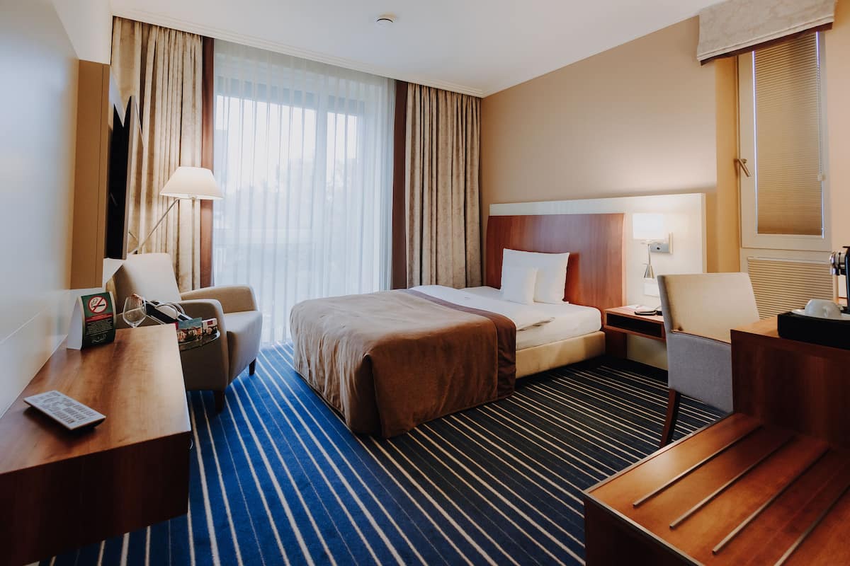 A hotel room with a bed, a desk, a chair, a nightstand, and a window with curtains.