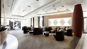 A spacious lobby with a modern design, featuring a curved reception desk, multiple seating areas with sofas and ottomans, and a large wall-mounted light fixture. - Lind Hotel (Rietberg)