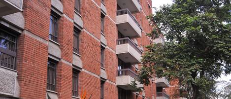 Un edificio de varios pisos de ladrillo con balcones, rodeado de abundante vegetación y plantas grandes.