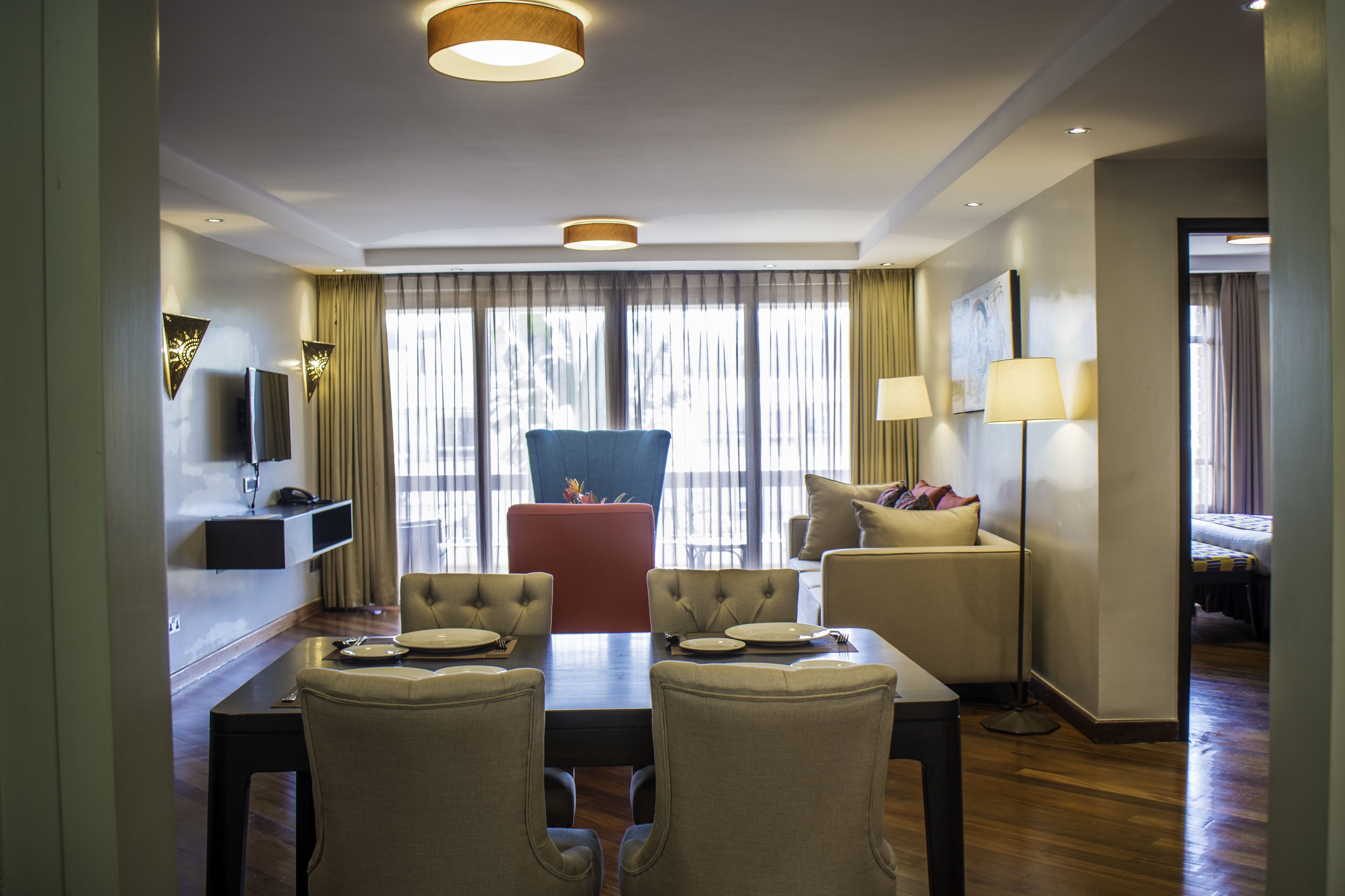 A hotel room with a dining area, a red chair, a sofa, a TV, and a lamp.