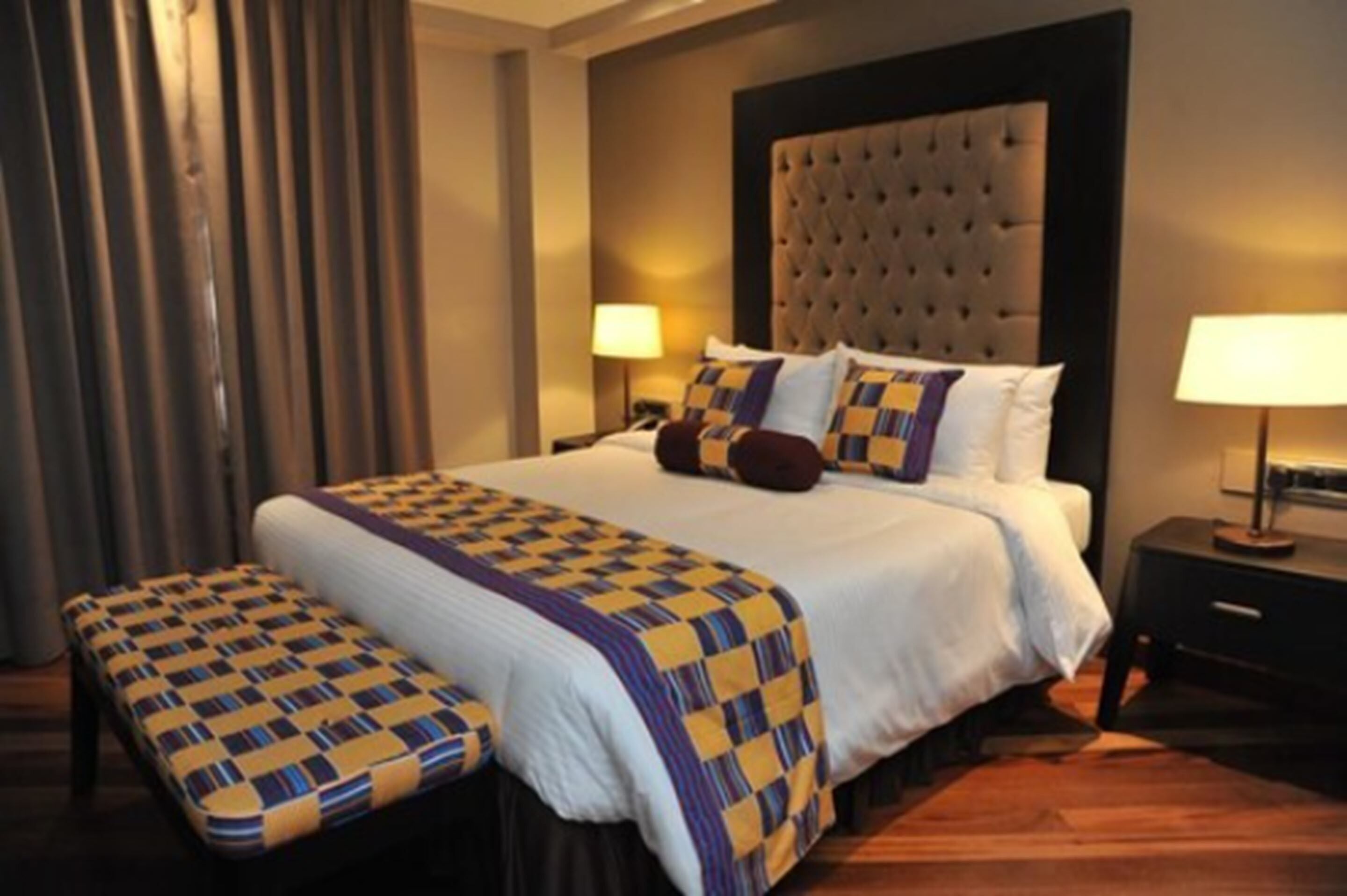 Una habitación de hotel con una cama grande, dos mesitas de noche, un banco y una ventana con cortinas.