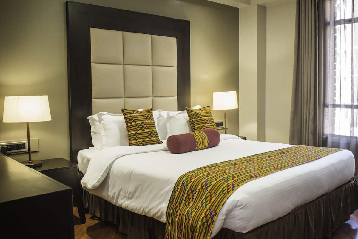 A hotel room with a large bed, two bedside lamps, a headboard, and a window with curtains.