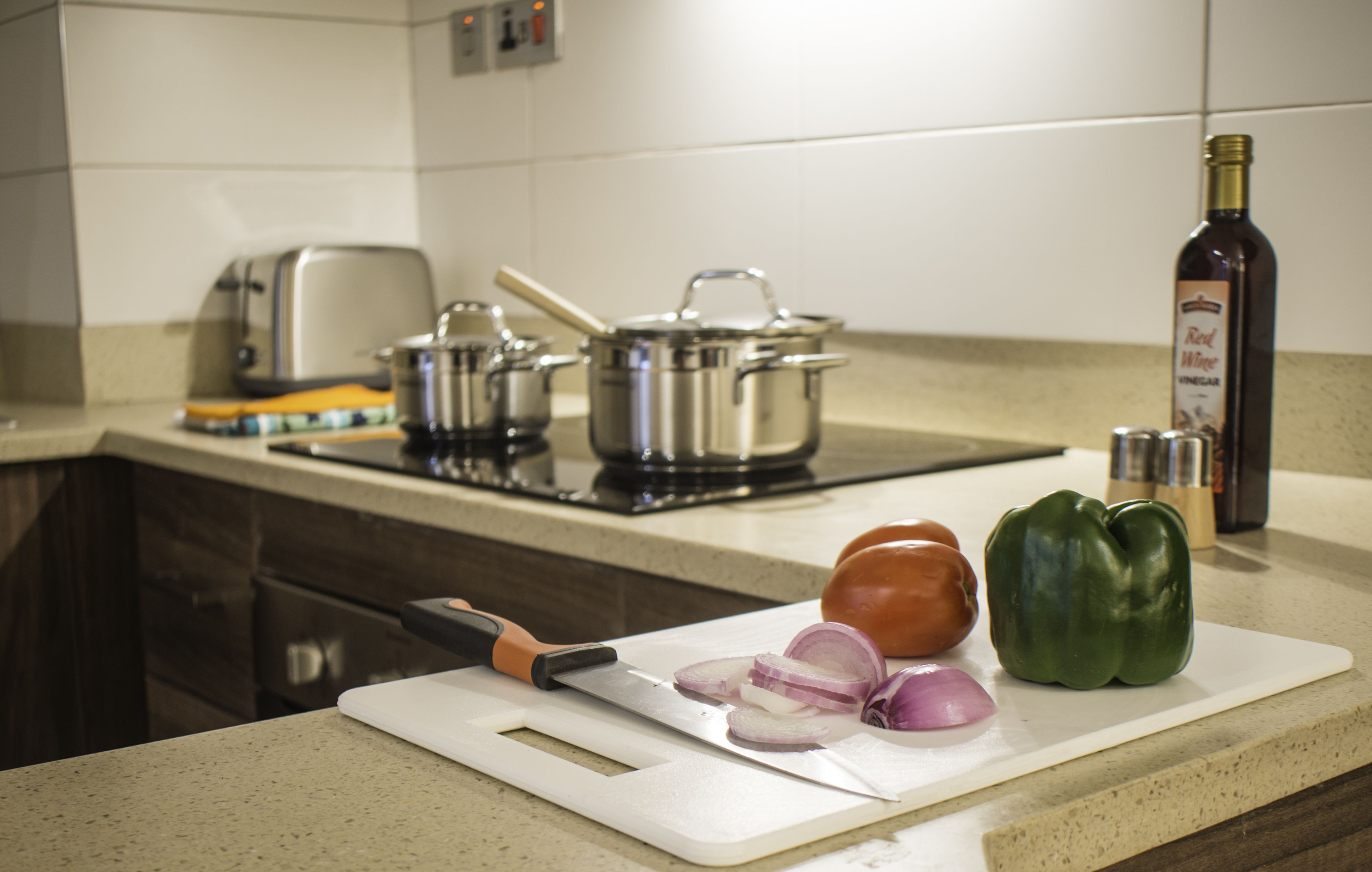 Un plan de travail de cuisine avec une planche à découper, un couteau, des oignons coupés en tranches, un poivron, une bouteille de vinaigre de vin rouge et des casseroles sur la cuisinière.
