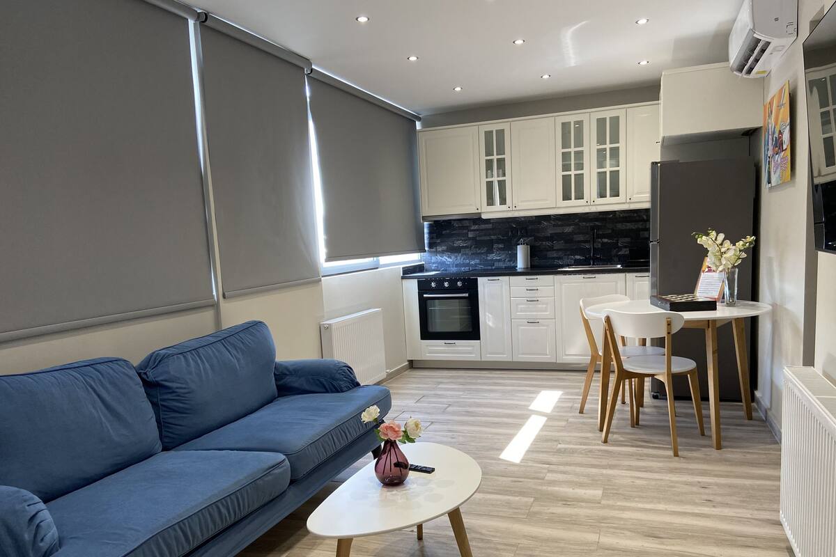 Una sala de estar moderna con un sofá azul, una mesita blanca con un florero y una cocina con armarios blancos y encimera negra.