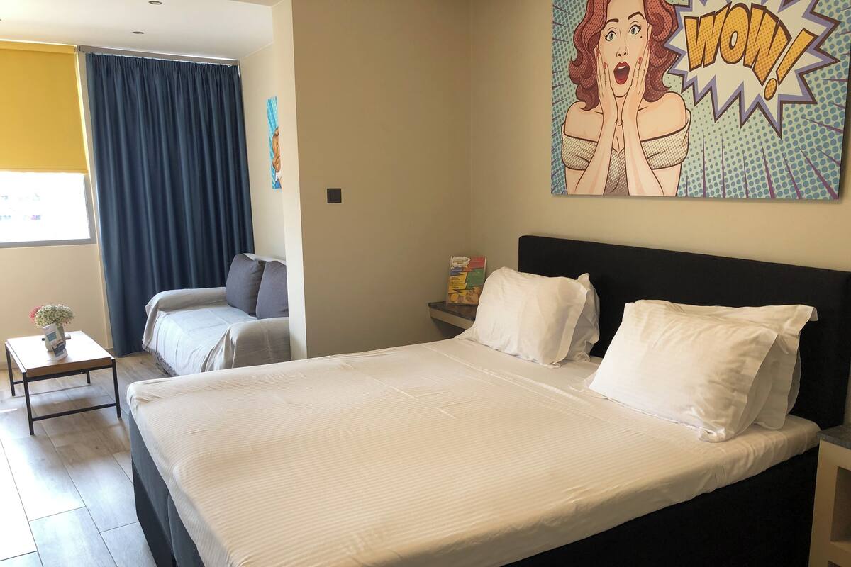 Habitación de hotel con una cama grande, un sofá más pequeño, una mesita de noche con lámpara y una ventana con cortinas.