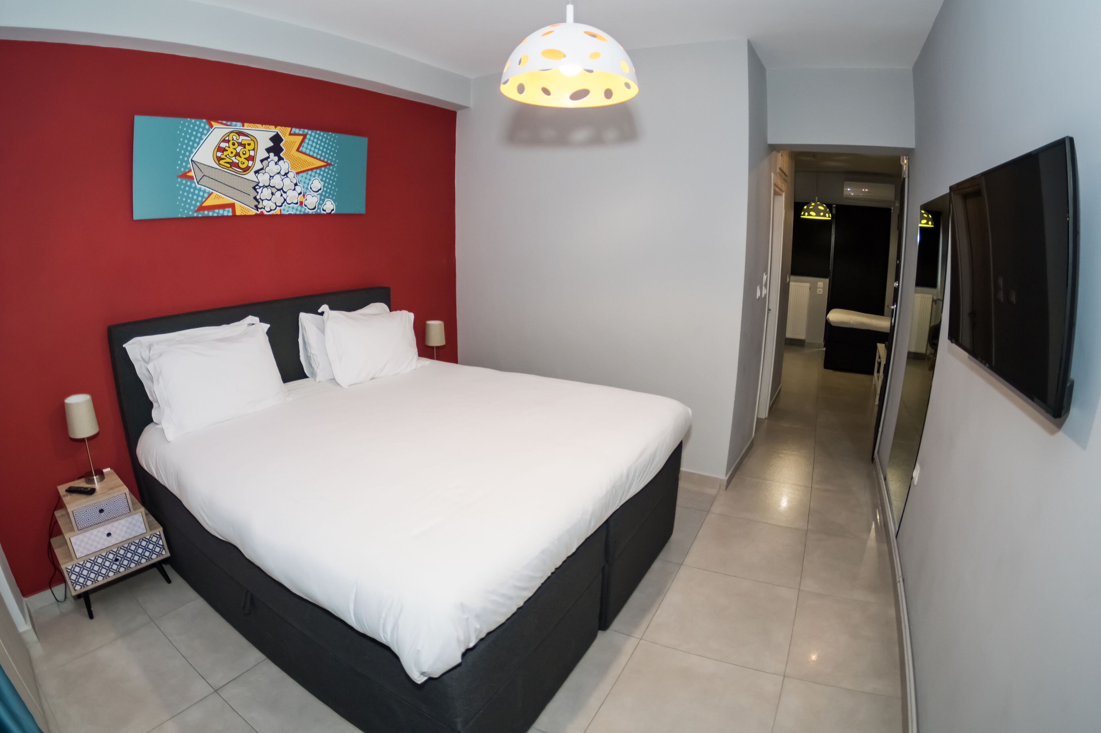 Una camera d'hotel con un letto grande, una parete rossa che fa da elemento decorativo, una lampada a sospensione, una televisione e un'apertura che dà su un'altra stanza.