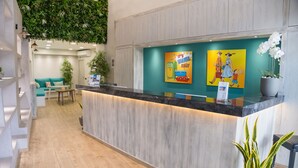 A reception area with a wooden reception desk, a teal wall with travel-themed artwork, and a green hanging plant. - Connect Studios & Apartments (Athens)