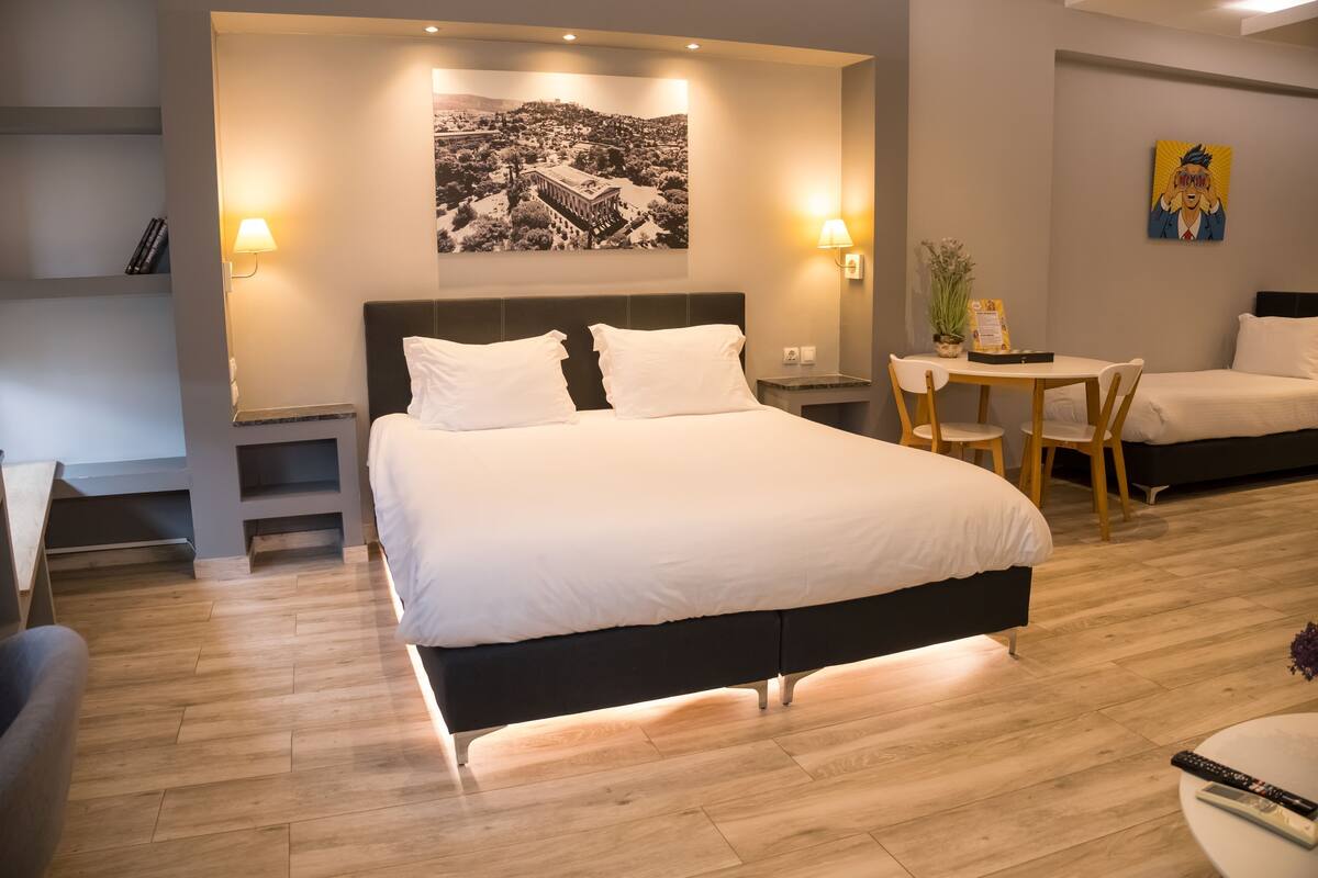 Habitación de hotel con una cama grande, mesitas de noche, un escritorio con sillas, una pequeña mesa con una planta y un cuadro colgado en la pared.