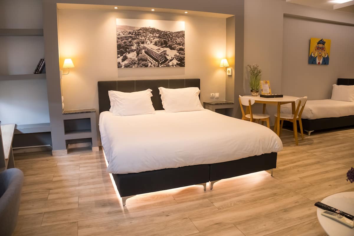 A hotel room with a large bed, bedside tables, a desk with chairs, a small table with a plant, and a wall-mounted painting.