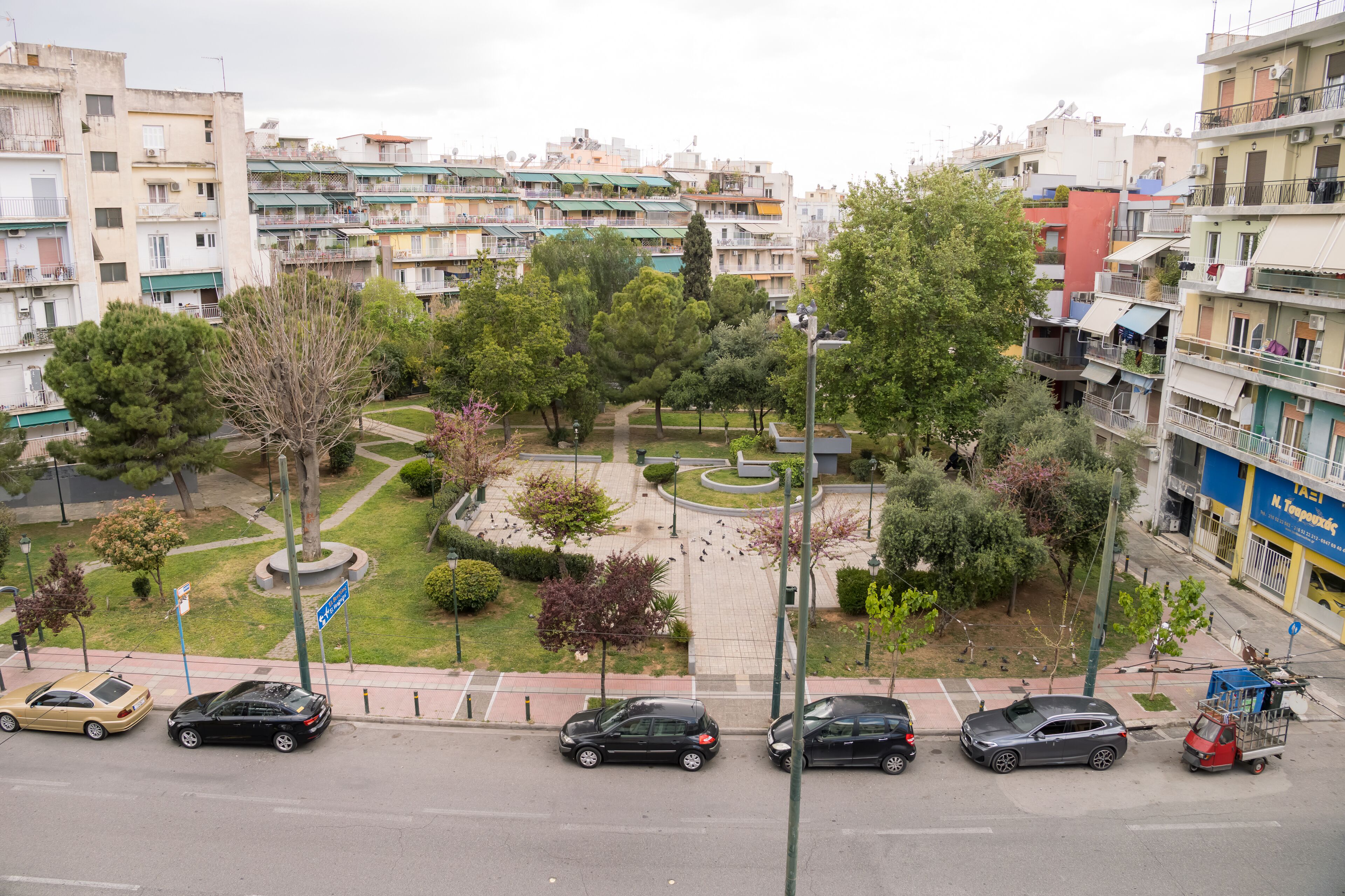 A residential area with multi-story buildings, a central park, and parked cars along the street.