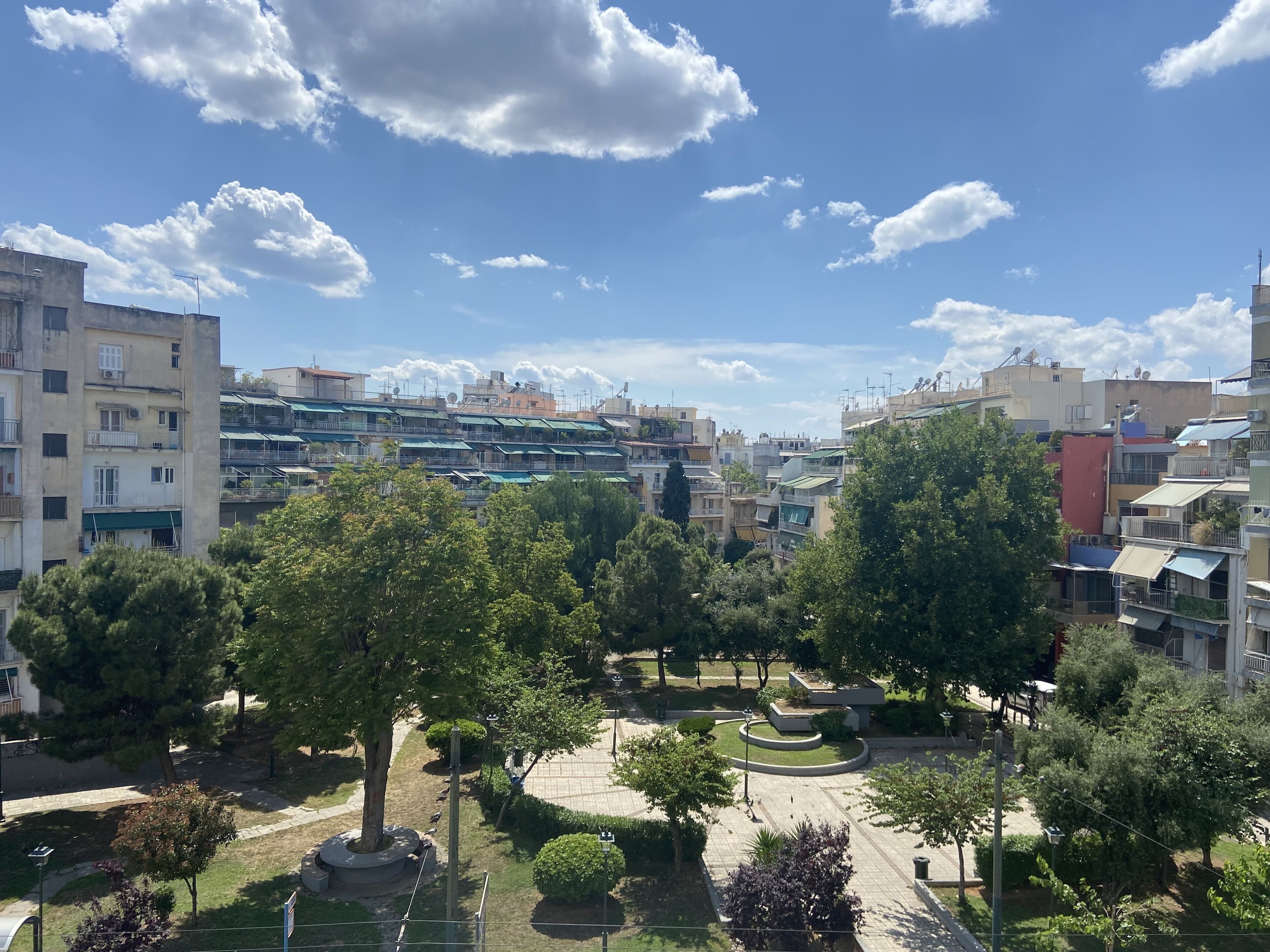 A residential area with multi-story buildings, a park with trees and a circular garden, and a clear blue sky with clouds.