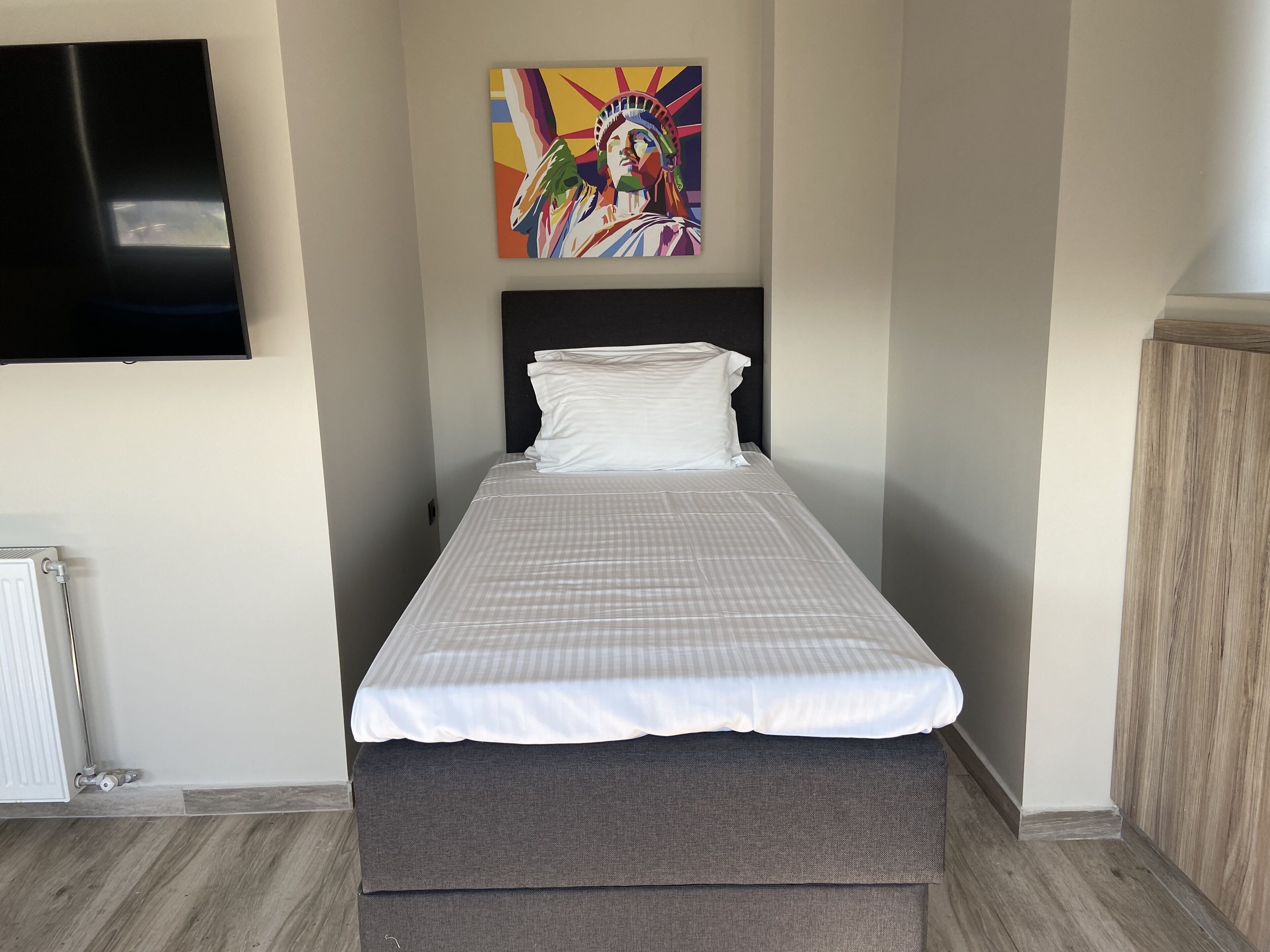 Un lit simple avec une couvre-lit à carreaux, un oreiller blanc et une tête de lit grise. Un tableau coloré représentant une figure est accroché au mur au-dessus du lit.