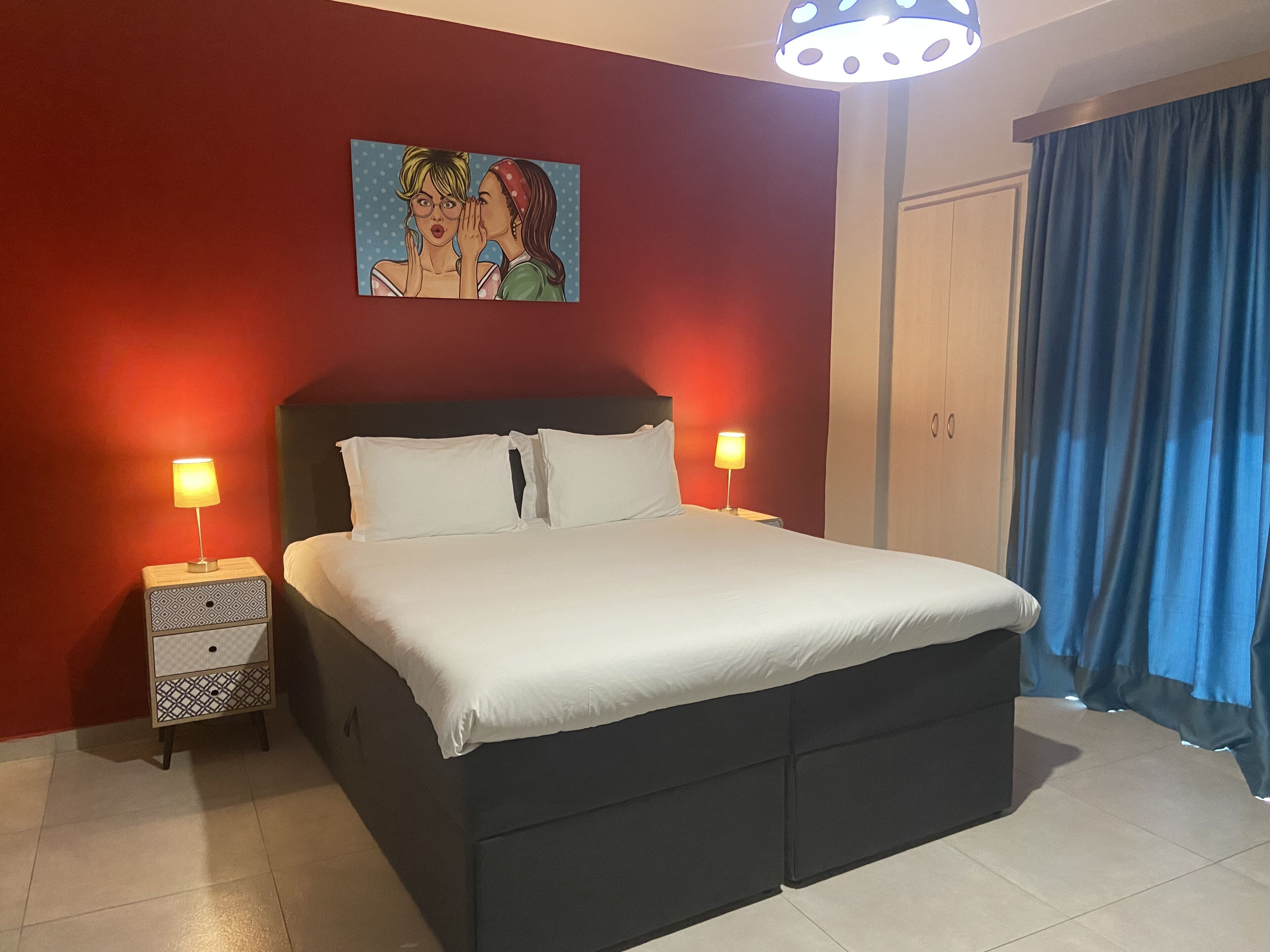 Un letto rifatto con lenzuola bianche, due comodini con lampade, un quadro sopra il letto e una tenda blu alla finestra.